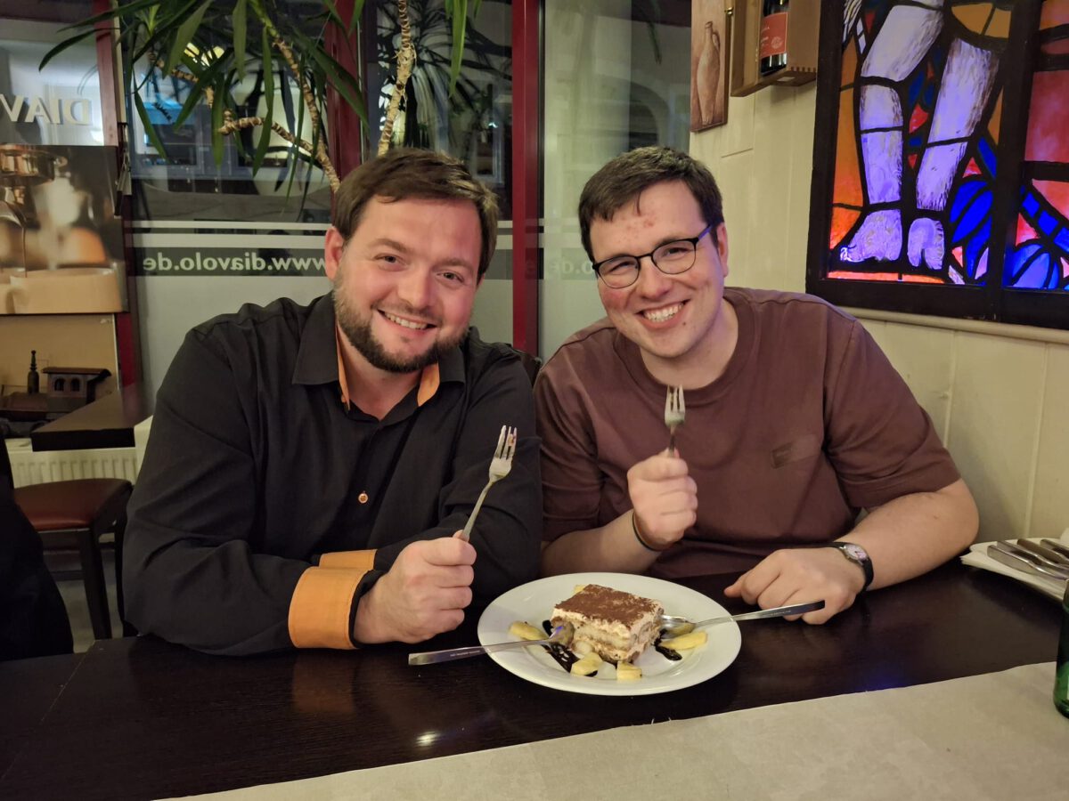 Robert und Karl sitzen fröhlich vor einem Tiramisu mit Gabeln in der Hand.